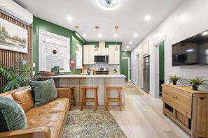 A modern kitchen with a central island, bar stools, a wooden cabinet, and a TV mounted on the wall. - Modern Verde • Chic 3BR • Easy NYC Commute (Jersey City)