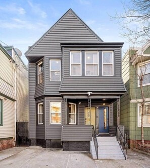 A two-story house with a gabled roof, a front porch with a black railing, and a blue door. - Modern Verde • Chic 3BR • Easy NYC Commute (Jersey City)