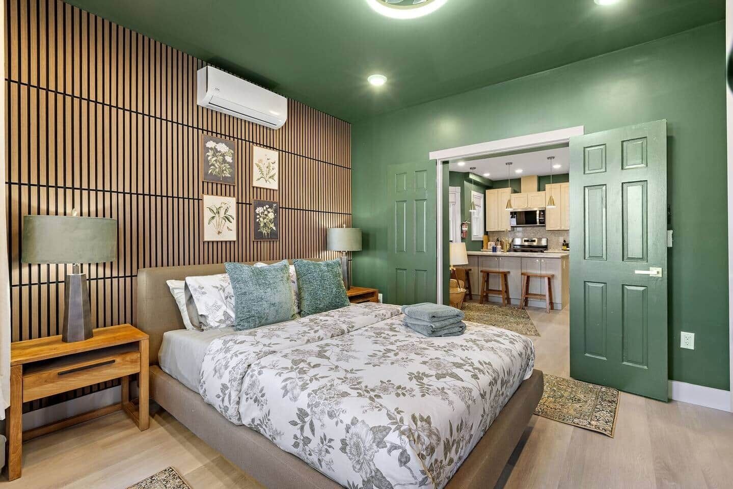 A bedroom with a bed, nightstands, a wall-mounted air conditioner, and a view into a kitchen area.