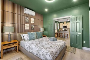 A bedroom with a bed, nightstands, a wall-mounted air conditioner, and a view into a kitchen area. - Modern Verde • Chic 3BR • Easy NYC Commute (Jersey City)