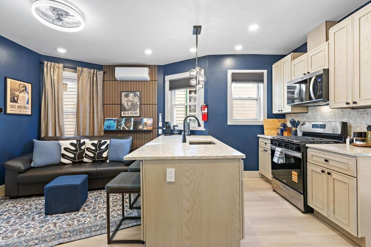 A modern kitchen with a central island, a dining area with a sofa and ottoman, and a living room with a blue wall and artwork.