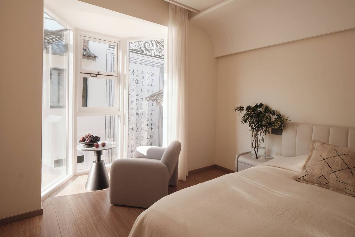 Una habitación de hotel con cama, una silla, una mesita con un florero y una ventana con cortinas.