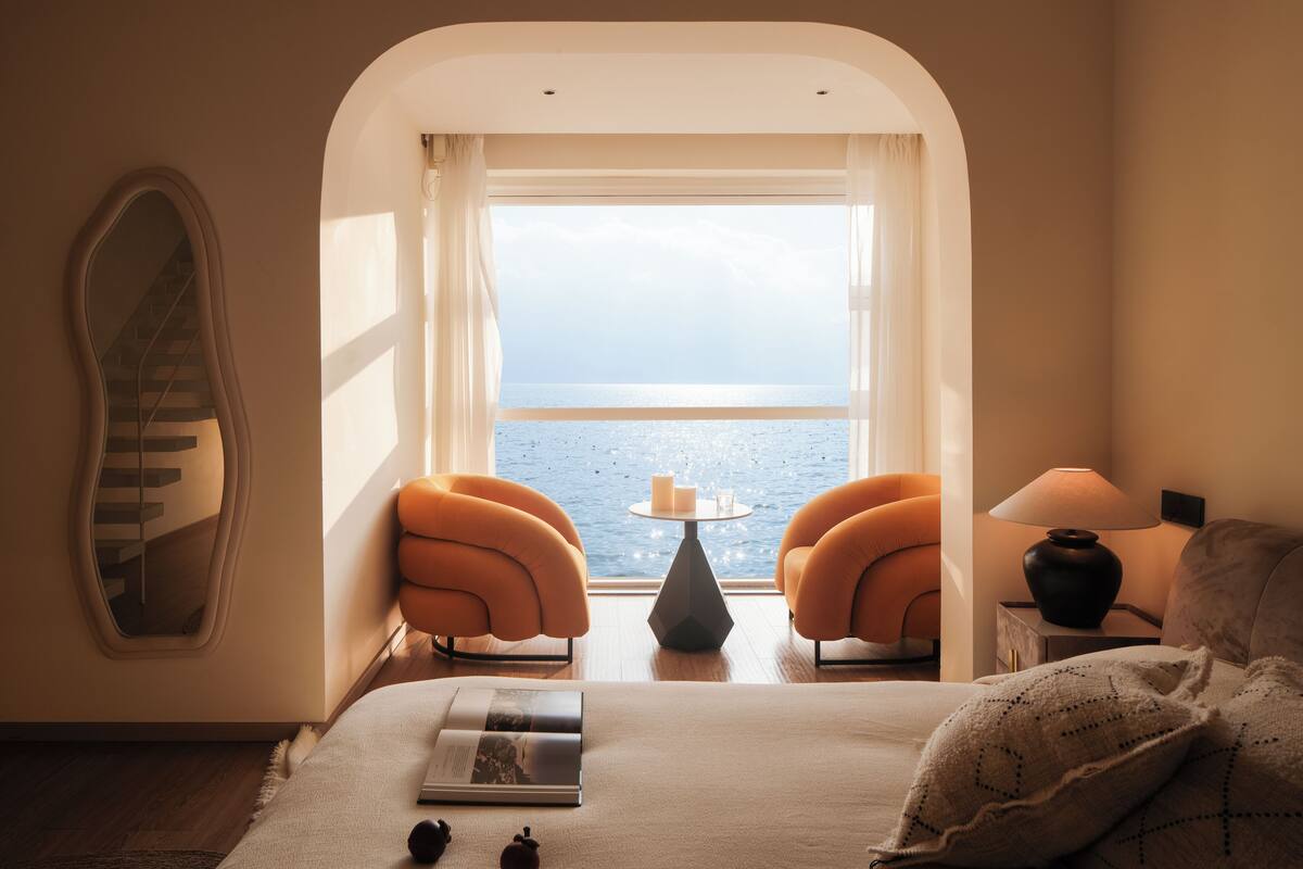 Un dormitorio con una cama, dos sillones naranjas, una mesita con una vela y vistas al mar a través de una ventana.