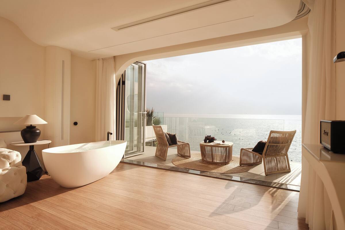 Habitación de hotel moderna con una bañera grande, sillones de mimbre, una mesa redonda y vista al mar.