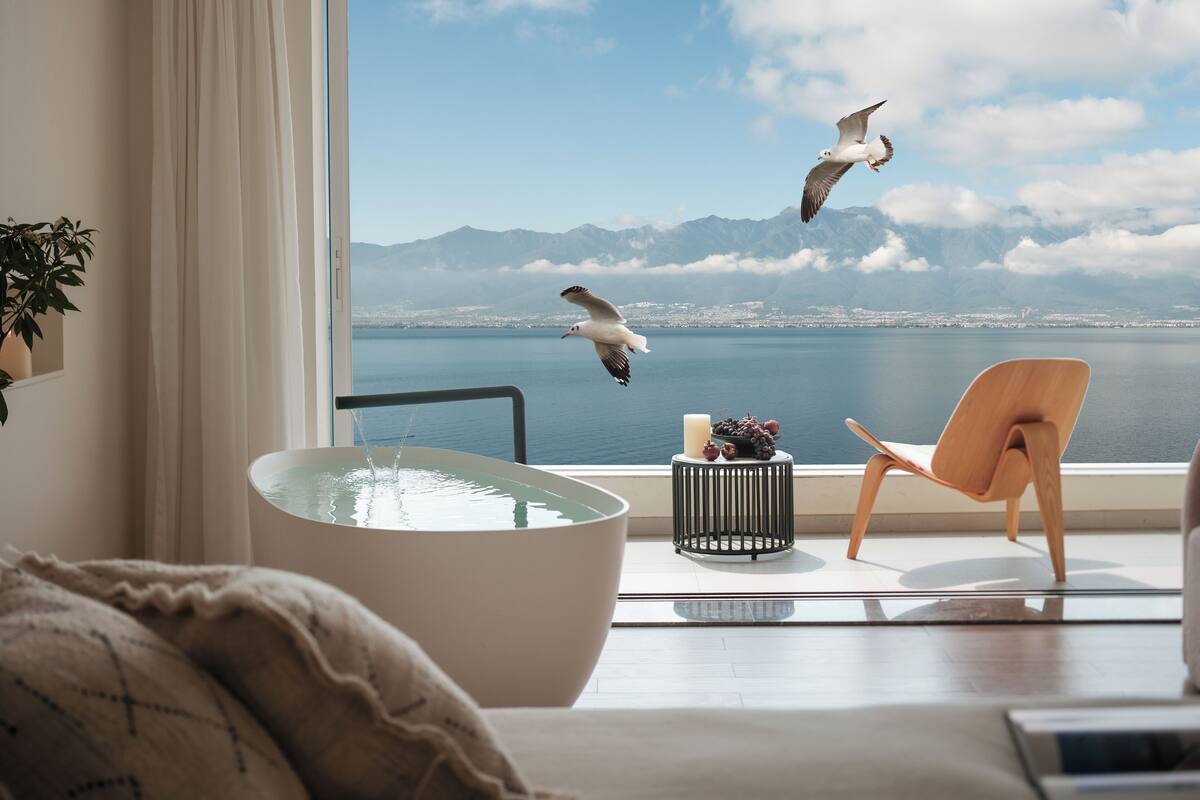 Habitación con amplia ventana que da a un cuerpo de agua y montañas. Hay una silla moderna y una mesa con una vela y uvas.