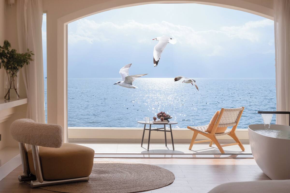 Una habitación moderna con un amplio ventanal con vistas al mar, que cuenta con una zona de estar cómoda con una silla y una mesa, y una bañera.