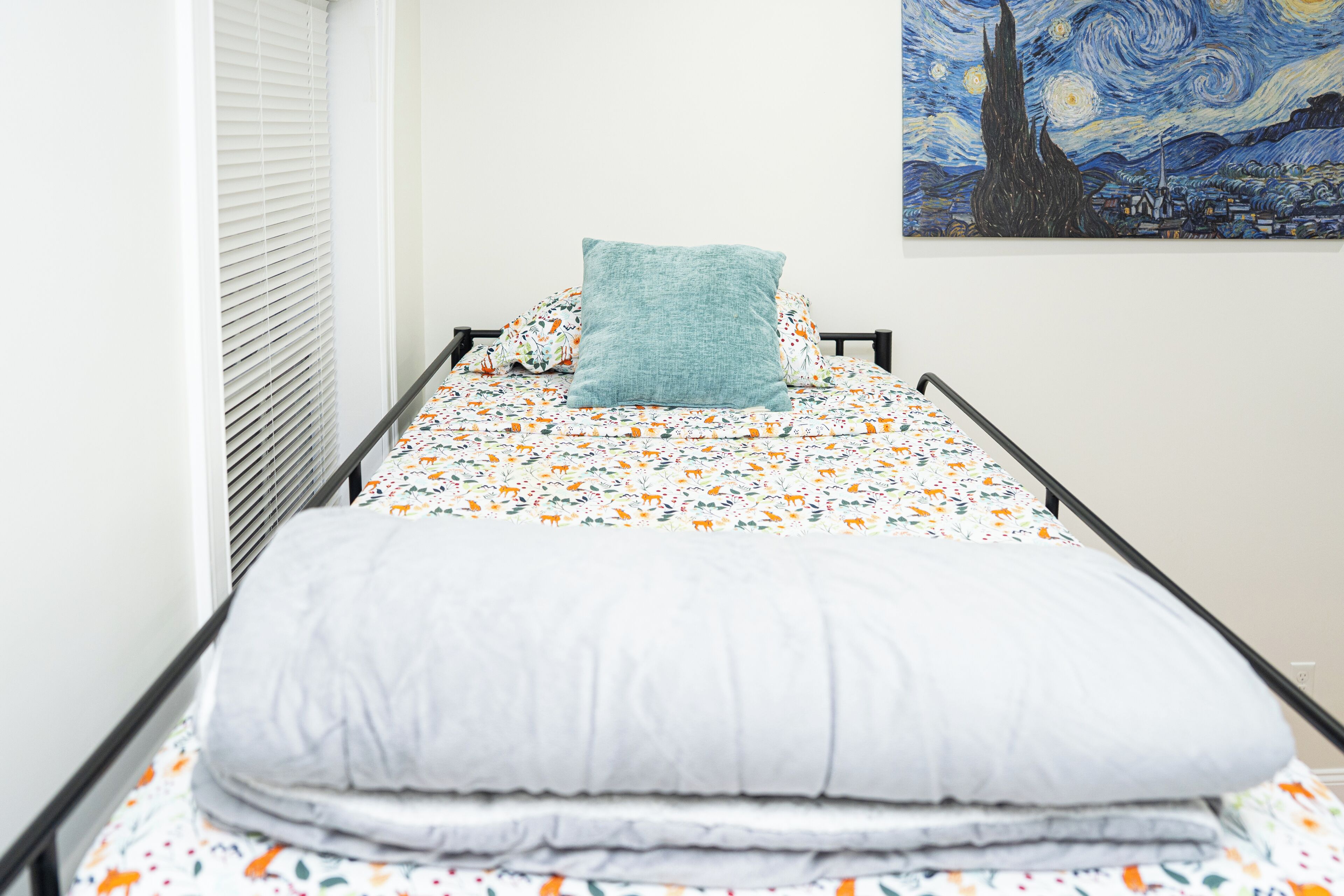 Una habitación pequeña con cama individual, cubierta con una colcha estampada de flores, un almohárga azul turquesa y una colcha blanca. Se observa un cuadro con un cielo nocturno lleno de estrellas en la pared.