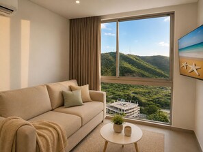 Ein Zimmer mit einem beigefarbenen Sofa, einem kleinen runden Tisch mit einer Pflanze und einer Tasse, einem großen Fenster mit Blick auf Grünflächen und ein Gebäude, sowie einem Strandbild an der Wand.