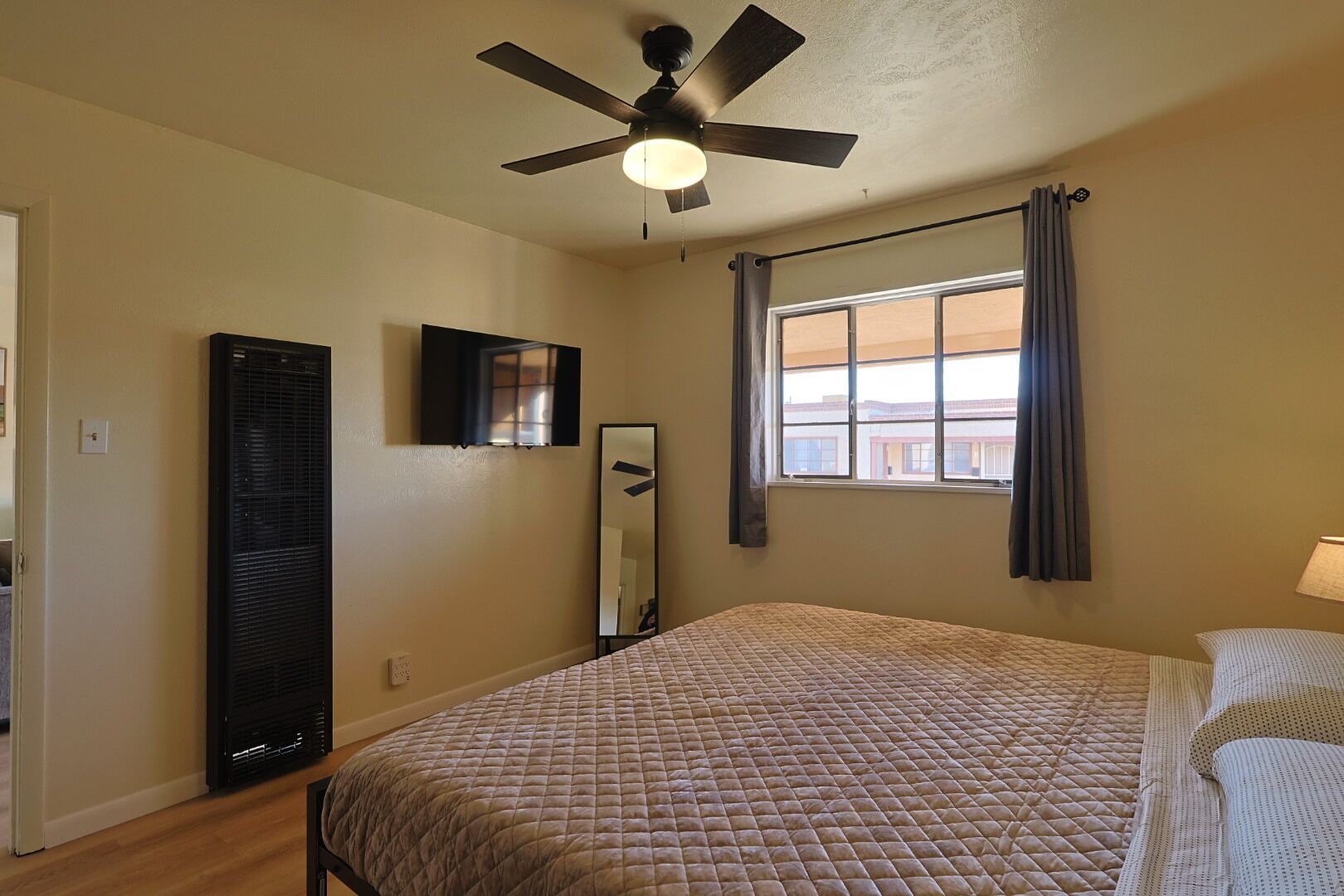 Un dormitorio con cama, ventilador de techo, ventana con cortinas, espejo instalado en la pared y un calentador.