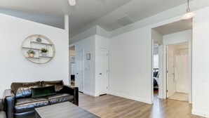 A spacious living room with a dark leather sofa, a coffee table, and a wall-mounted shelf with plants. - Meridian Gem / 1 King+ 2 Queens / Central location (Meridian)