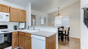 A modern kitchen with wooden cabinets, a white countertop, a built-in dishwasher, and a dining area with a chandelier. - Meridian Gem / 1 King+ 2 Queens / Central location (Meridian)