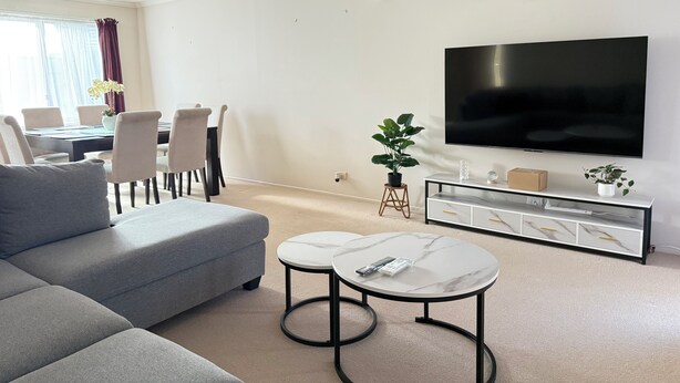 Een moderne woonkamer met een grijze hoekbank, een marmeren salontafel, een eethoek met een tafel en stoelen, een flatscreen tv aan de muur en een potplant.