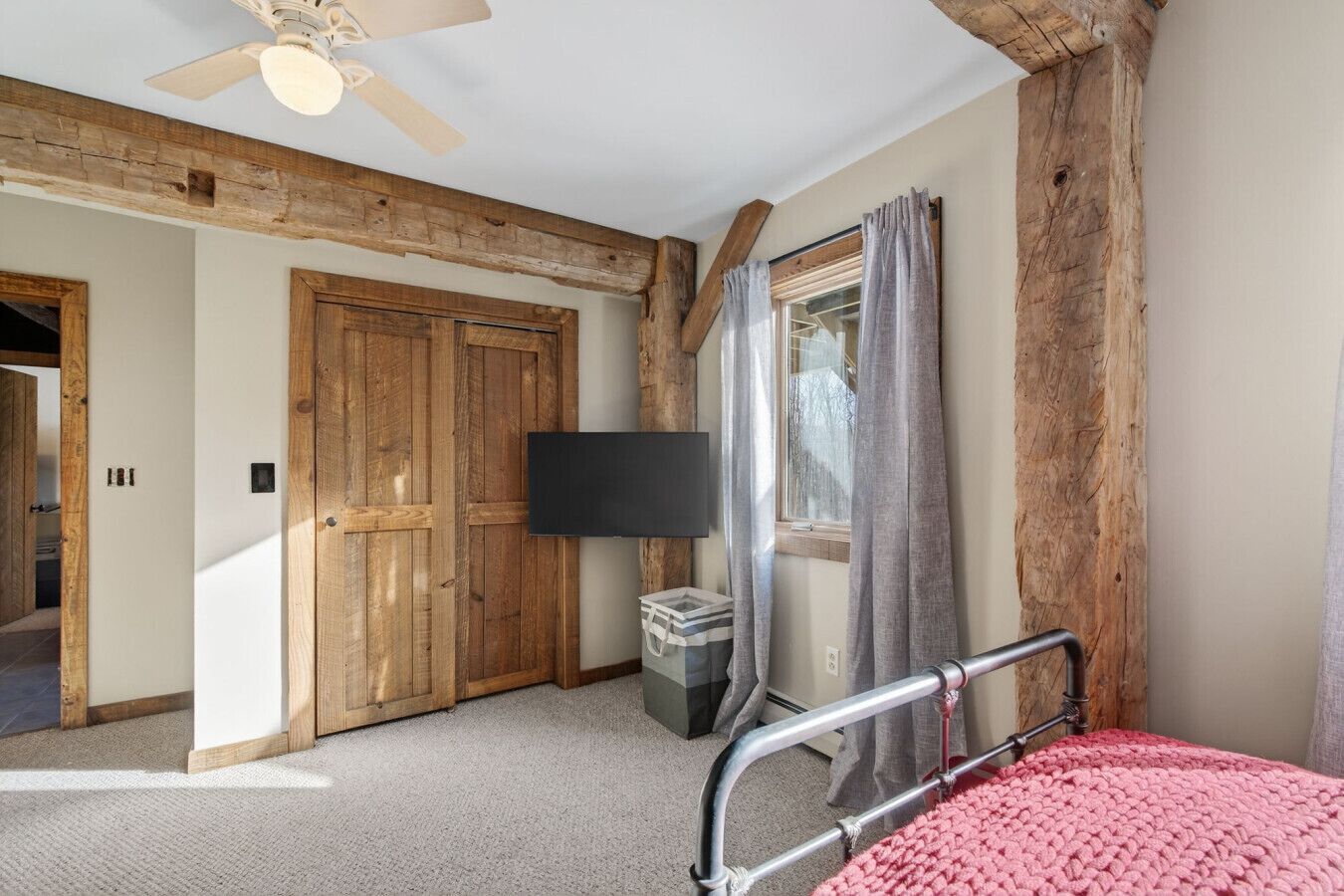A bedroom with a wooden ceiling, a bed with a metal frame, a TV mounted on the wall, and a window with curtains.