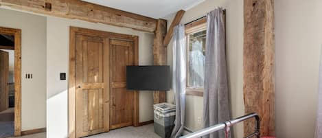 A bedroom with a wooden ceiling, a bed with a metal frame, a TV mounted on the wall, and a window with curtains.