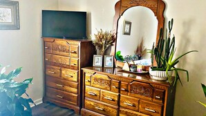 Une chambre avec une commode en bois, un téléviseur à écran plat, un grand miroir et un lit recouvert d’une couvre-lit à motifs floraux.