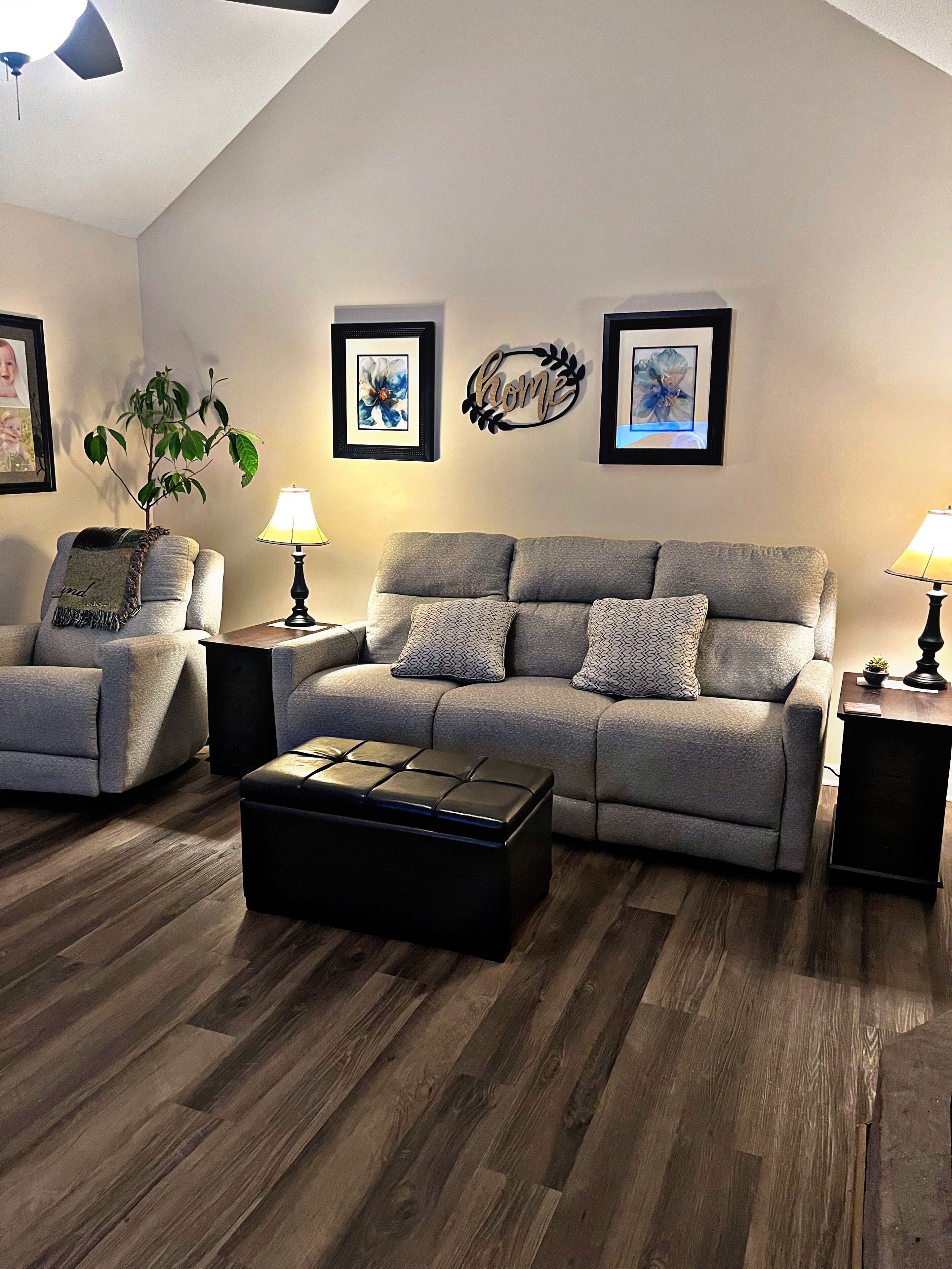 A living room with a grey sofa, a matching armchair, a black ottoman, and a wooden floor. There are two wall-mounted lamps and a framed artwork above the sofa.