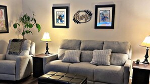 Un salon avec un canapé gris, un fauteuil assorti, un pouf noir et un plancher en bois. Deux lampes murales et une œuvre d’art encadr ée sont disposées au-dessus du canapé.
