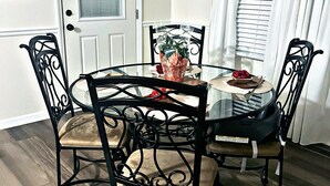 Un coin repas comprenant une table ronde en verre, quatre chaises en métal noir, une porte blanche, une fenêtre avec des rideaux blancs et un luminaire fixé au mur.