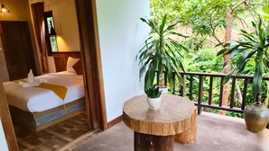 Una habitación con cama, una mesita con una planta y un balcón con plantas en macetas.