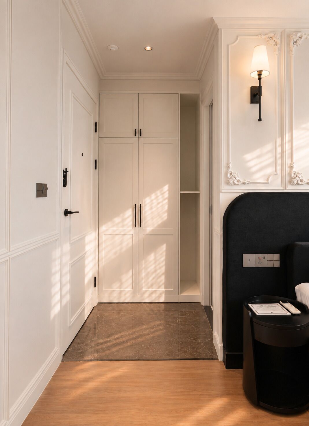 A hotel room with a wooden floor, a black headboard, a bedside table with a lamp, and a wardrobe with a mirror.