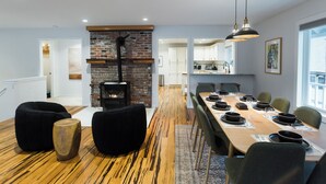Un espace repas moderne avec une longue table en bois dressée pour un repas, une cheminée en briques et une cuisine en arrière-plan.