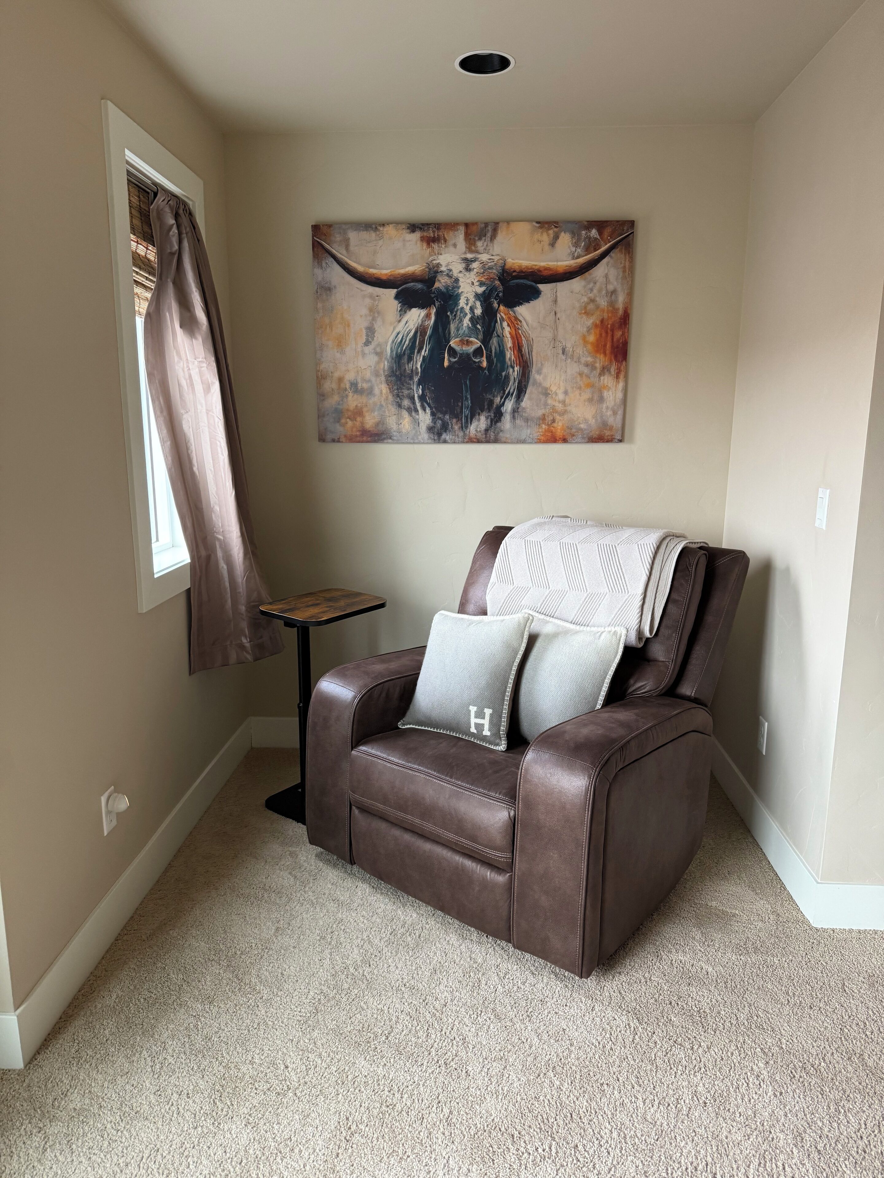 Een bruine lederen fauteuil met een deken en kussens, een raam met gordijnen, en een schilderij van een stier aan de muur.