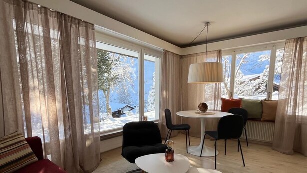 A modern living room with a red sofa, two white round tables, and a dining area with chairs. Large windows offer a snowy mountain view. - Haus Rupicapra by Interhome (Arosa)