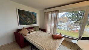 A bedroom with a bed, a red sofa, a painting, a window with a view of trees and mountains, and a small table. - Haus Rupicapra by Interhome (Arosa)