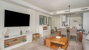 Ein modernes Wohnzimmer mit Flachbildfernseher, Couchtisch aus Holz und einem Essbereich mit Stühlen und Tisch.