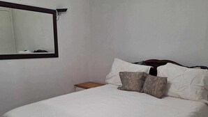 Ein ordentlich bezogenes Bett mit weißen Leinen und mehreren Kissen, ein Nachttisch aus Holz und ein Spiegel an der Wand.