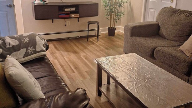 Ein Wohnzimmer mit braunem Ledersofa, einem gemusterten Sessel, einem Holztisch, einem an der Wand montierten Flachbildfernseher und einer Topfpflanze.