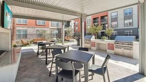 An outdoor patio area with tables and chairs, a barbecue grill, and a view of residential buildings. - Brand-New Build-Luxury 2BR Retreat-Resort Pool -Travel Nurse Friendly-Buckeye AZ (Buckeye)