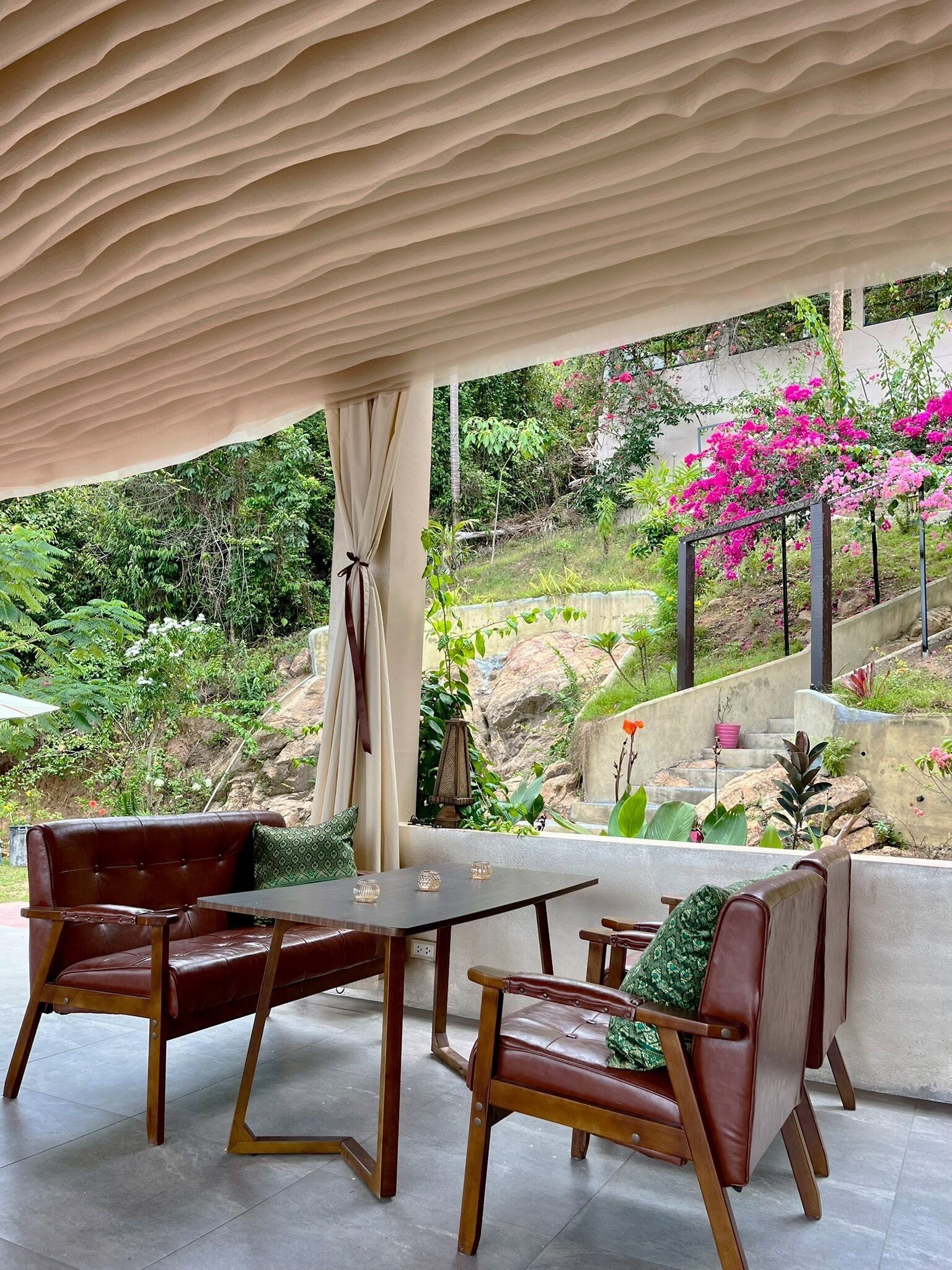 Un espacio al aire libre con mesa y sillas, una zona cubierta con cortina y vistas a un jardín lleno de flores rosadas y follaje.