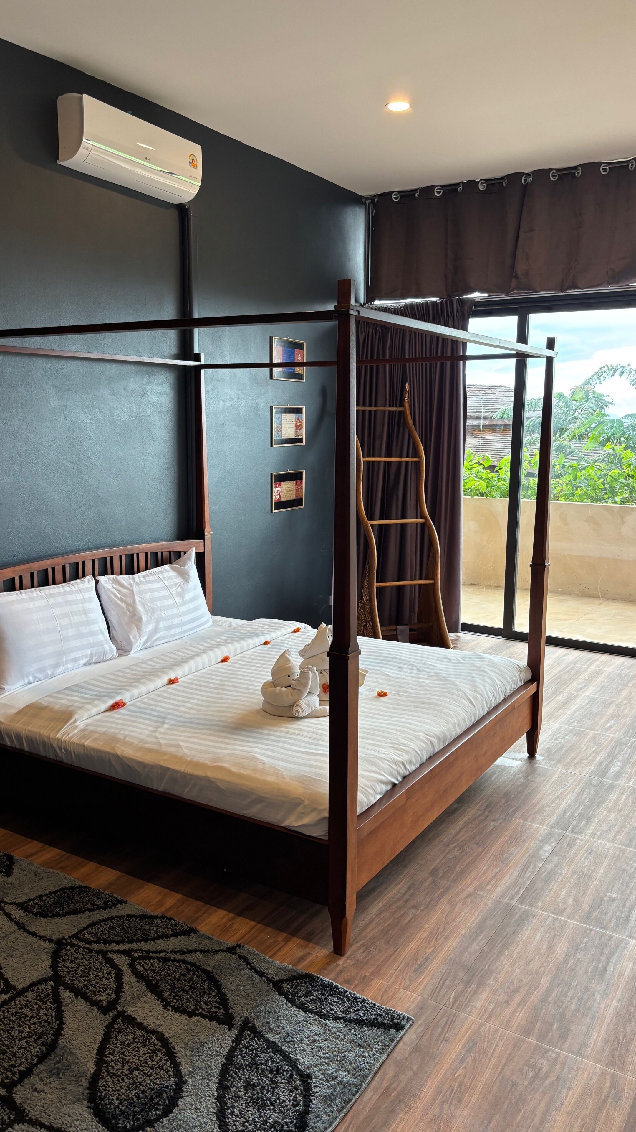 Habitación de hotel con una cama balinesa de madera, un ventanal con cortinas y aire acondicionado de pared, con vistas a zonas verdes.