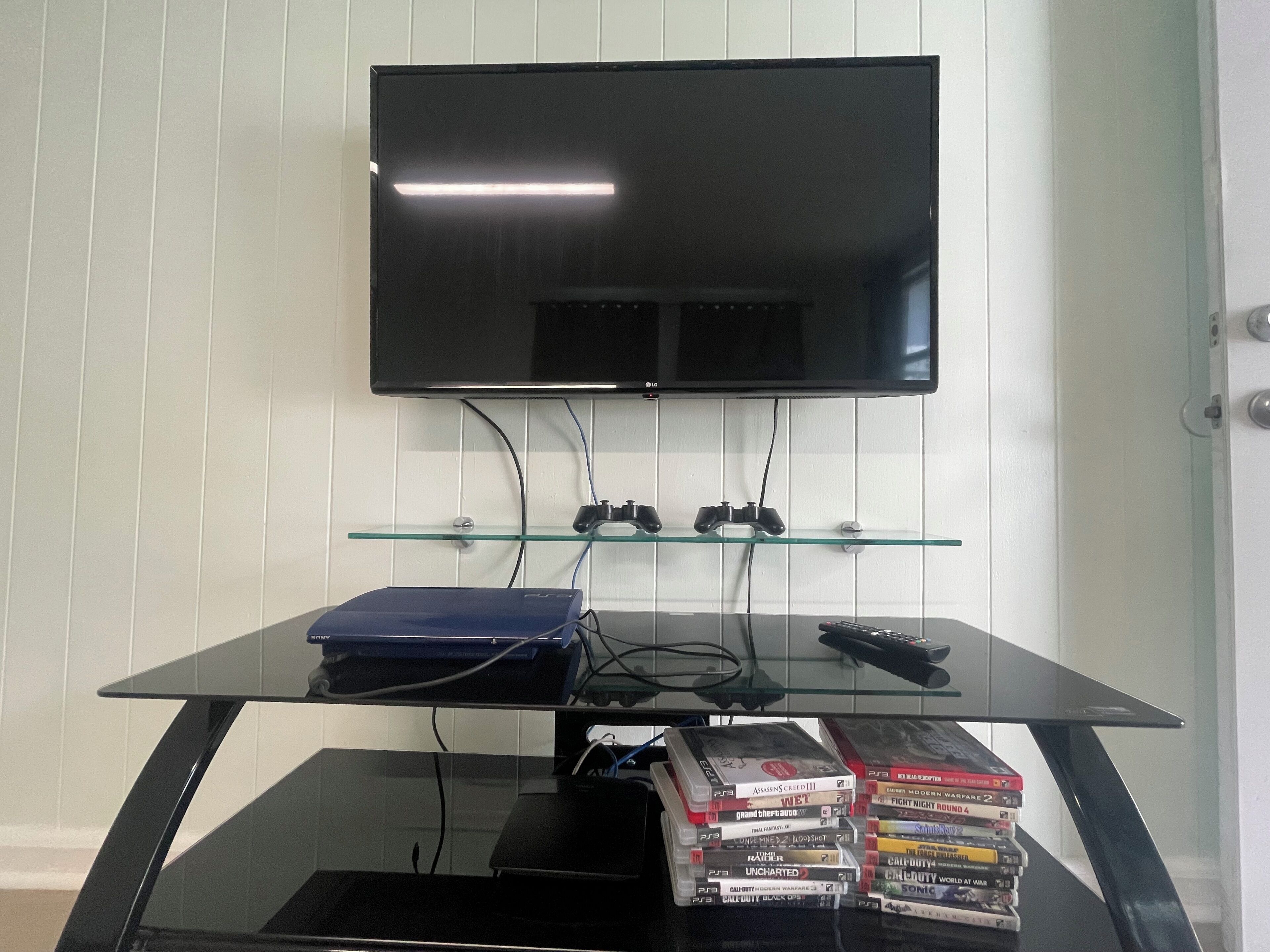Un téléviseur à écran plat fixé au mur, au-dessus d’une table basse en verre, sur laquelle se trouvent une télécommande et des manettes de jeu.