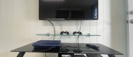 Un téléviseur à écran plat fixé au mur, au-dessus d’une table basse en verre, sur laquelle se trouvent une télécommande et des manettes de jeu.
