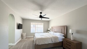 Une chambre à coucher comprenant un lit, une table de chevet, une lampe, un ventilateur de plafond, un téléviseur fixé au mur et une fenêtre avec des stores.
