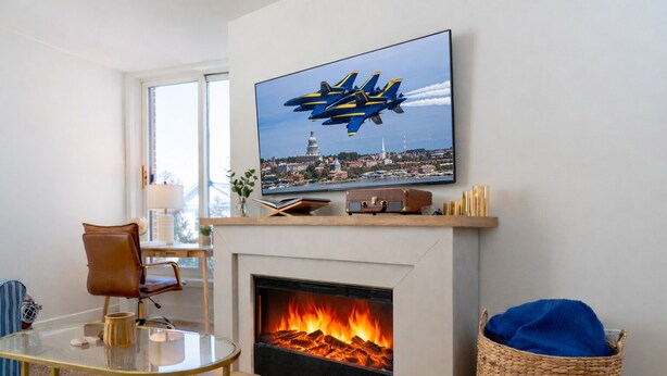 Ett vardagsrum med en öppen spis, en tv som hänger ovanför och visar ett flygplan, och ett soffbord i glas.