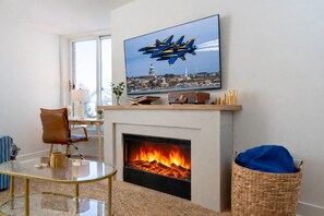 A living room with a fireplace, a television mounted above it displaying an airplane, and a glass coffee table.