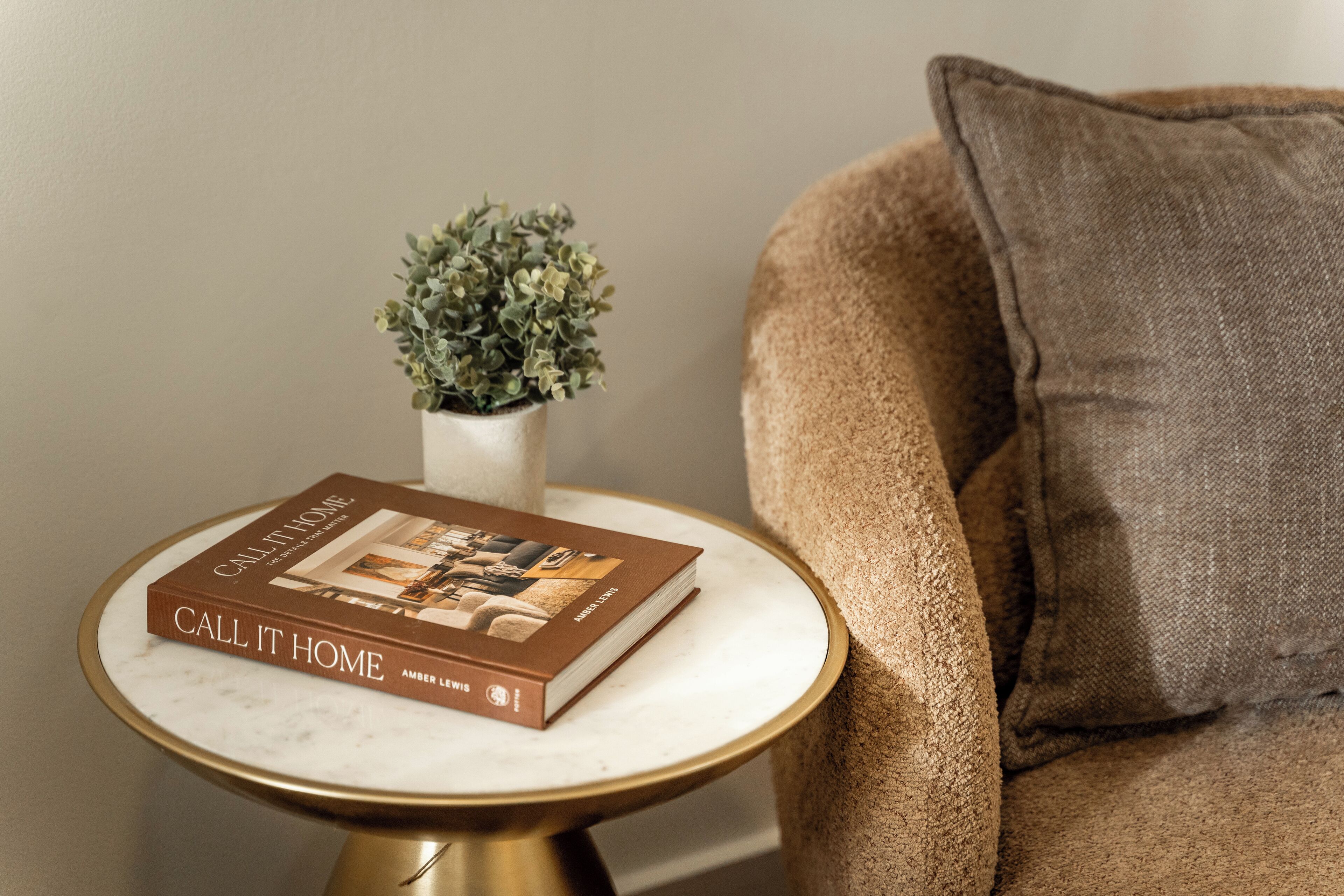 A book titled "Call It Home" on a round table with a gold base, next to a beige couch with a brown pillow.