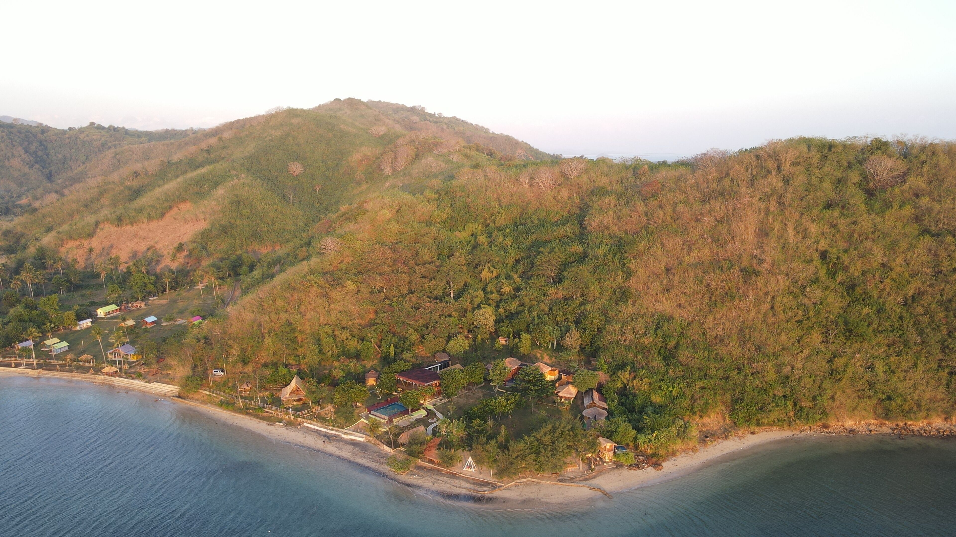An aerial view of a coastal area with a beach and hills. There are several buildings, likely accommodations, scattered along the coastline.