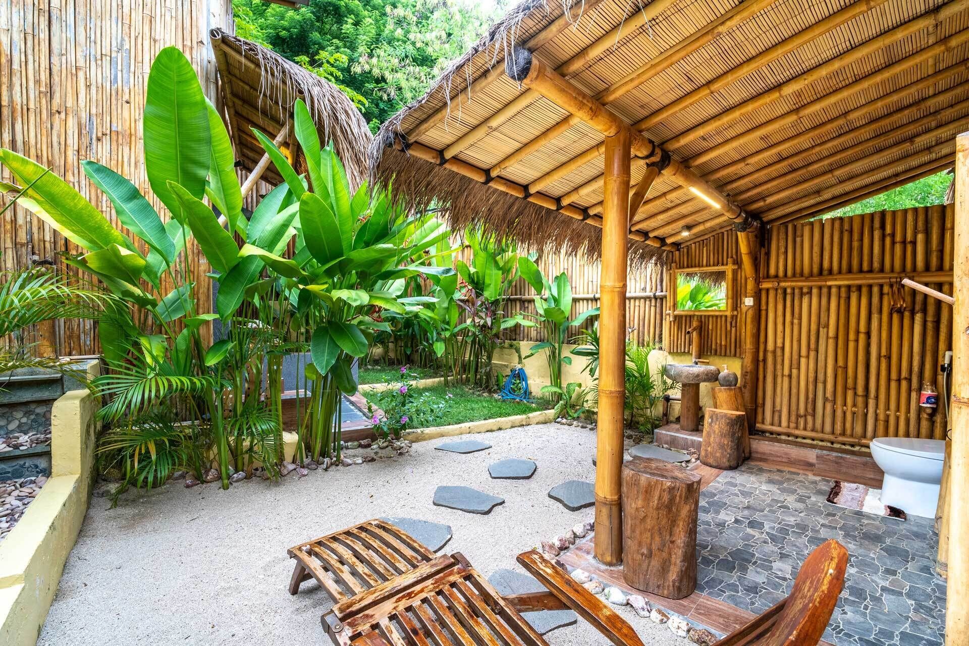 A rustic outdoor area with a thatched roof, stone seating, and lush greenery.