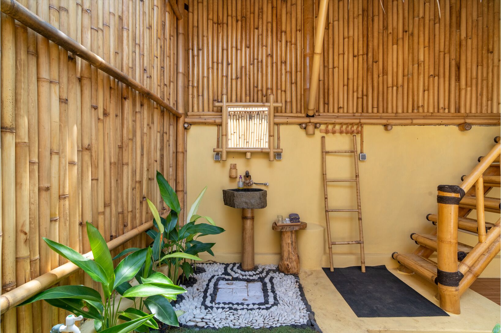 A bamboo-walled room with a stone sink, a wooden ladder, and a small table with a plant.