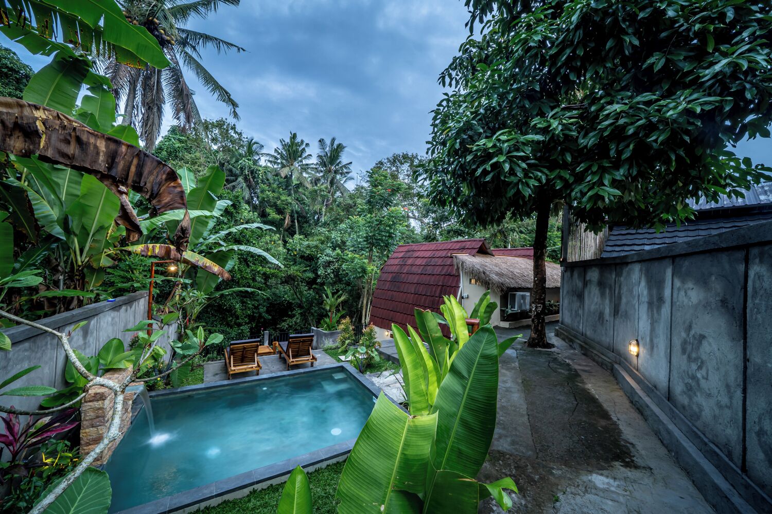 Un área de piscina privada con sillas de descanso, rodeada de exuberante vegetación tropical y un edificio con techo de paja.