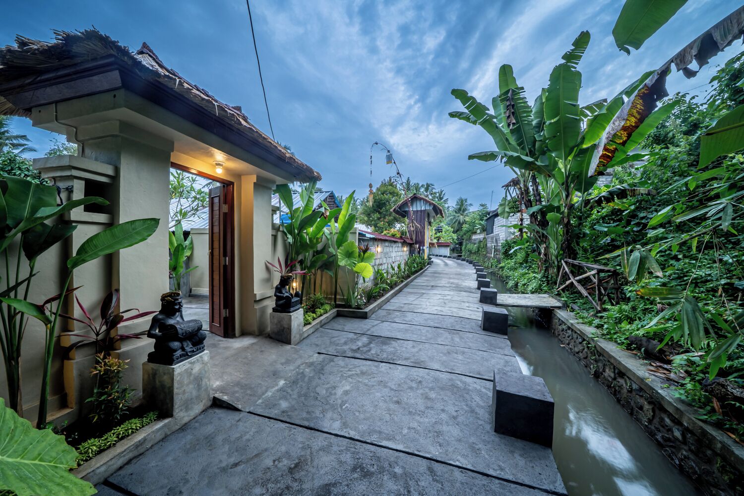 A pathway leading to a house with a thatched roof, surrounded by lush greenery and a small stream.