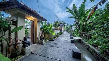 A pathway leading to a house with a thatched roof, surrounded by lush greenery and a small stream.