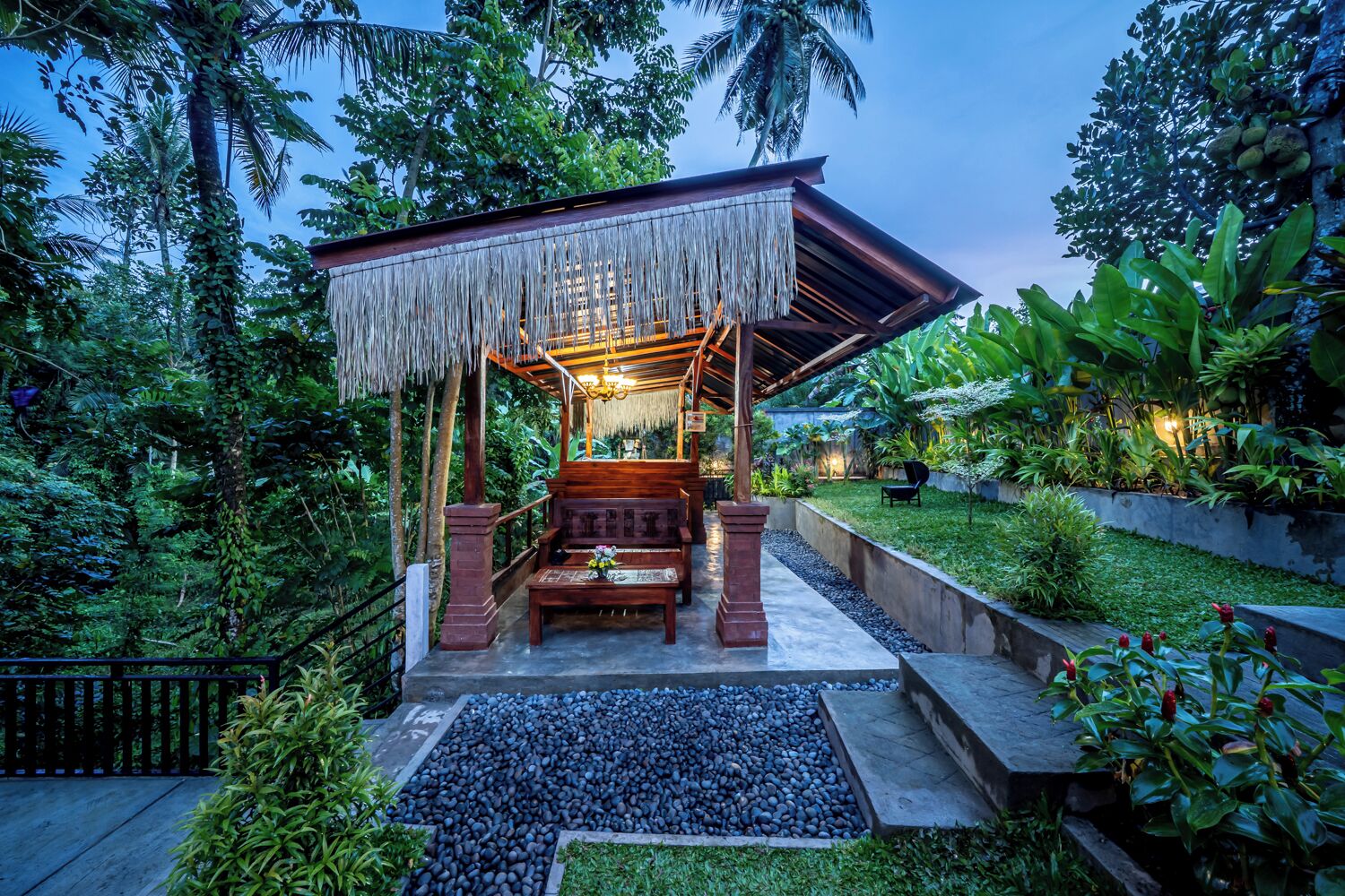 A wooden gazebo with a thatched roof, surrounded by lush greenery and a well-maintained garden.