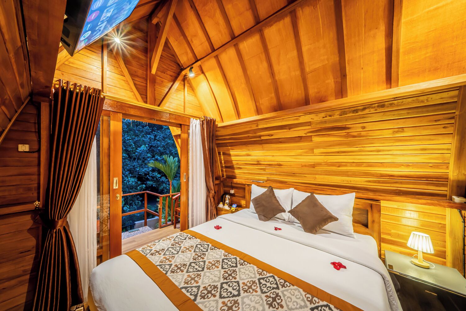 Un dormitorio de cabaña de madera con cama, cortinas, una mesita de noche y vistas al exterior.