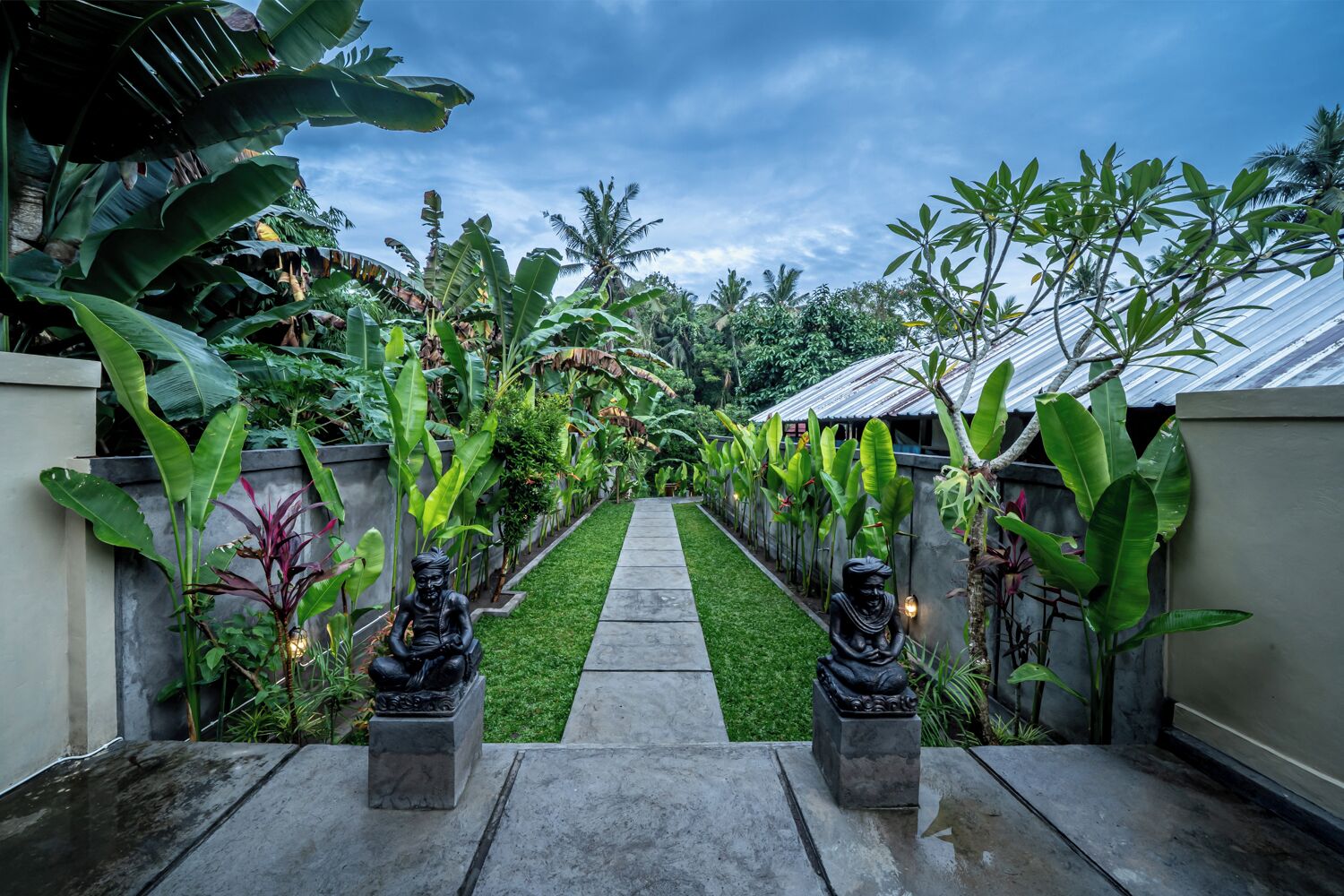 A pathway flanked by statues and lush tropical plants leading to a building with a metal roof.