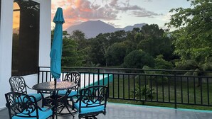 A balcony with a table and chairs, a blue umbrella, and a view of mountains and trees.
